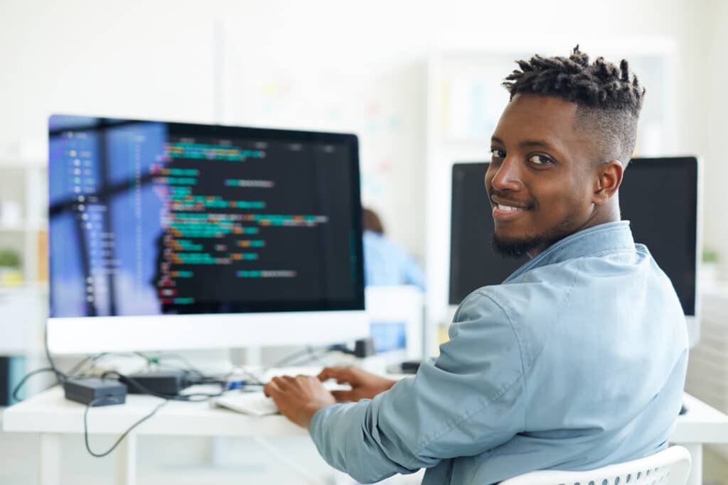 Happy guy looking at you while sitting by workplace and working with software